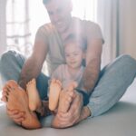 A man and a child sit on a bed, facing each other with their legs extended and feet touching—perhaps playfully comparing foot size or checking for signs of wide feet. The man is smiling, and both are wearing casual clothes. Sunlight filters through the window in the background.